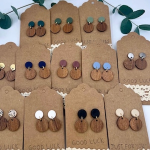 May include: A collection of wooden earrings with various colored tops, displayed on brown tags with the text "GOOD LUCK" and "JUST FOR YOU". The earrings have a circular wooden base and a smaller, colored circle on top. Some earrings have a gold-colored post.