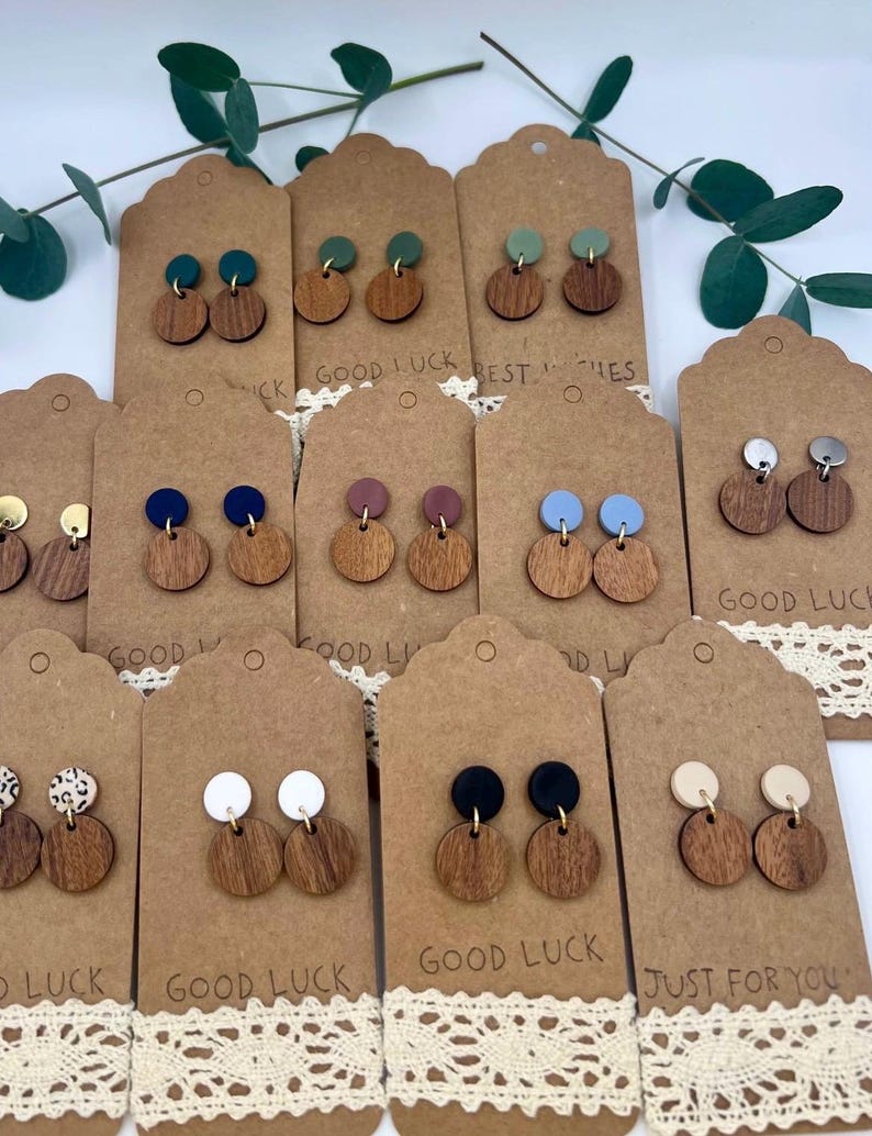 May include: A collection of wooden earrings with various colored tops, displayed on brown tags with the text "GOOD LUCK" and "JUST FOR YOU". The earrings have a circular wooden base and a smaller, colored circle on top. Some earrings have a gold-colored post.