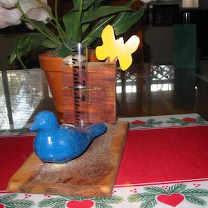 Puede incluir: Una figurita de pájaro azul se sienta sobre una base de madera con un tubo de vidrio transparente y un bloque de madera. Un recorte de mariposa amarilla está unido al bloque de madera. La base está sobre un mantel rojo y blanco con un borde verde.