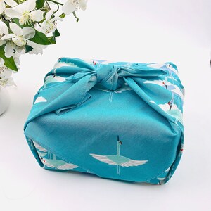 May include: A square, turquoise fabric wrap tied in a knot, featuring a repeating pattern of white cranes with outstretched wings against a cloudy sky. The fabric is wrapped around an unseen object. White flowers are in the background.