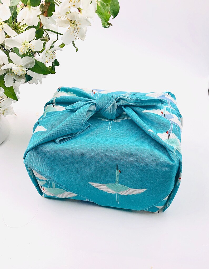 May include: A square, turquoise fabric wrap tied in a knot, featuring a repeating pattern of white cranes with outstretched wings against a cloudy sky. The fabric is wrapped around an unseen object. White flowers are in the background.