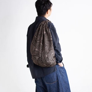Può includere: Una borsa con coulisse a fantasia marrone e nera è drappeggiata sulla spalla di una persona. La borsa presenta un motivo a onde ripetuto. La persona indossa una camicia blu scuro con bottoni e jeans.