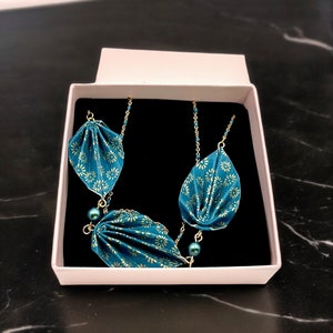 May include: A teal necklace with a floral pattern, presented in a white box with a black interior. The necklace features three folded leaf-shaped pendants connected by delicate gold chains and adorned with small teal beads.