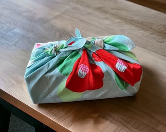 May include: A square package wrapped in a light blue cloth with a floral pattern. The design features large red flowers with pink centers and green leaves. The cloth is tied at the top with a matching blue ribbon.