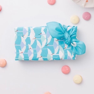 May include: A wrapped gift with a blue and white crane pattern, tied with a light blue bow. The gift is surrounded by pastel-colored macarons on a white background. The overall aesthetic is clean and elegant.