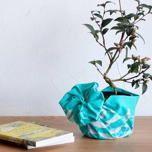 May include: A small potted plant with green leaves and pink buds sits in a turquoise fabric pot cover with a white and blue wave pattern. A book with a yellow cover rests on a wooden surface.