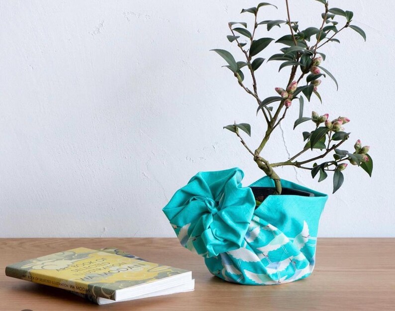 May include: A small potted plant with green leaves and pink buds sits in a turquoise fabric pot cover with a white and blue wave pattern. A book with a yellow cover rests on a wooden surface.