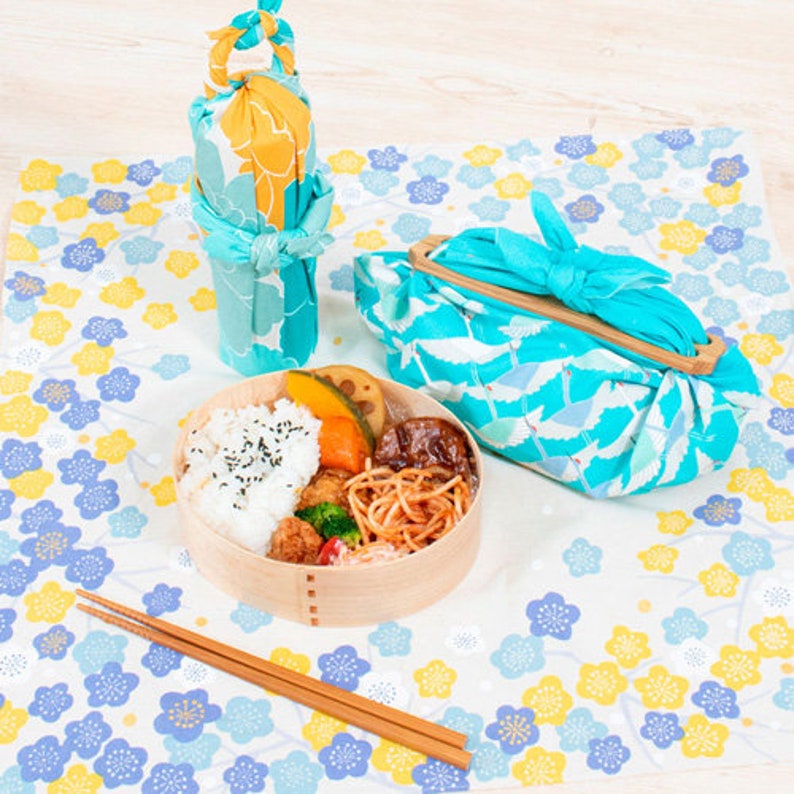 May include: A wooden bento box filled with rice, vegetables, and noodles, accompanied by chopsticks. A blue and yellow floral patterned cloth and matching fabric wraps a bottle and a lunch bag. The scene is set on a white surface with a floral pattern.