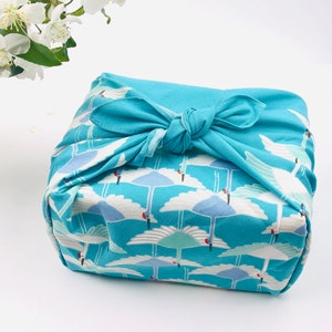 May include: A square package wrapped in a teal fabric with a repeating pattern of white cranes against a blue sky. The fabric is tied with a matching teal bow. White flowers are in the background.