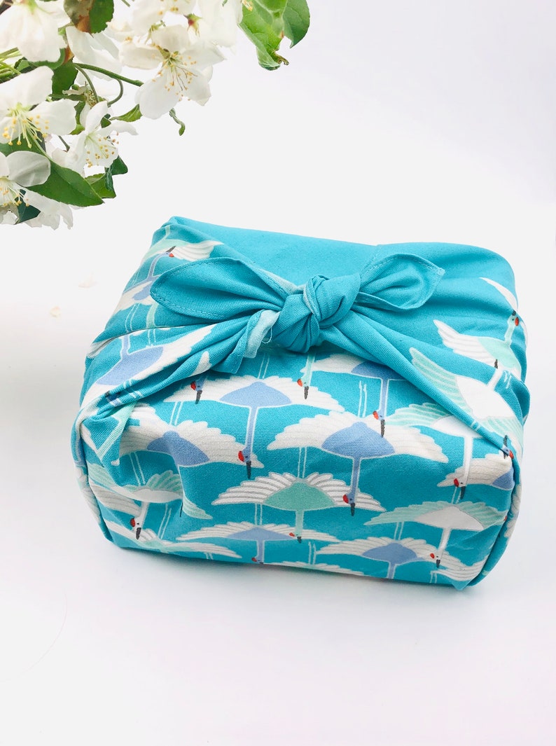 May include: A square package wrapped in a teal fabric with a repeating pattern of white cranes against a blue sky. The fabric is tied with a matching teal bow. White flowers are in the background.