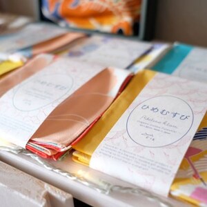 May include: A display of folded fabric items, each wrapped with a paper band featuring Japanese text and a circular design. The fabrics are in various colors, including peach, yellow, and blue, with some displaying patterns.