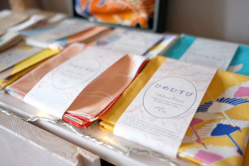 May include: A display of folded fabric items, each wrapped with a paper band featuring Japanese text and a circular design. The fabrics are in various colors, including peach, yellow, and blue, with some displaying patterns.