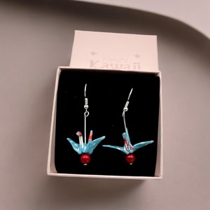 May include: A pair of origami crane earrings in a pink box. The earrings feature blue and red paper cranes with a red bead at the base. The box has the text "Tiny Kawaii" on the top.