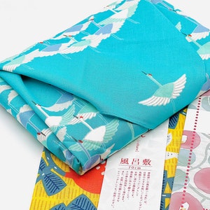 May include: Folded square fabric with a vibrant turquoise crane pattern. The fabric is folded over a yellow and blue patterned fabric, with a white label featuring Japanese text. The fabric measures 70cm.