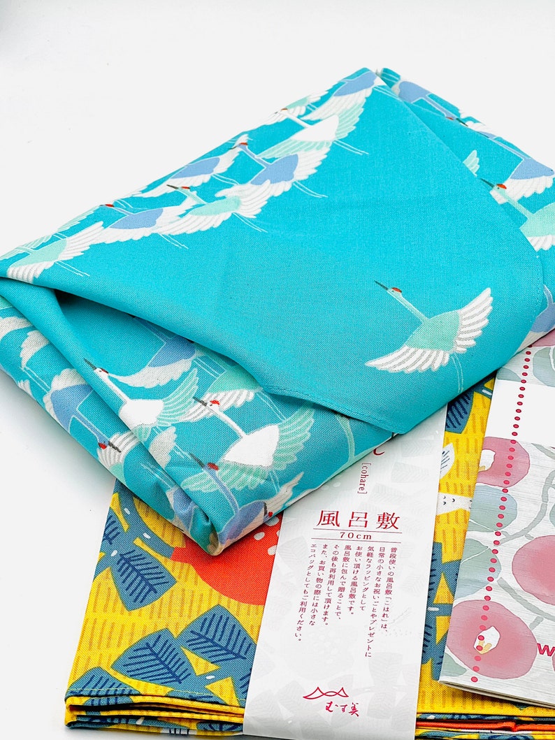 May include: Folded square fabric with a vibrant turquoise crane pattern. The fabric is folded over a yellow and blue patterned fabric, with a white label featuring Japanese text. The fabric measures 70cm.