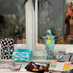 May include: Various fabric-wrapped gifts and items on a windowsill. The gifts are wrapped in patterned fabrics, including black and white, teal, and blue designs. A bottle is wrapped in a teal and yellow fabric. A black paper star hangs above.