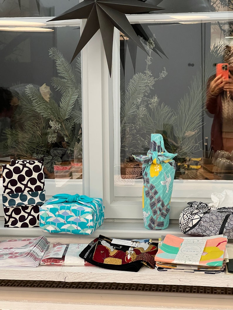 May include: Various fabric-wrapped gifts and items on a windowsill. The gifts are wrapped in patterned fabrics, including black and white, teal, and blue designs. A bottle is wrapped in a teal and yellow fabric. A black paper star hangs above.