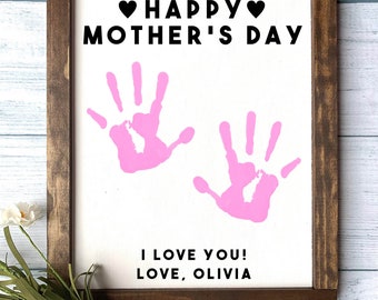 May include: A white wooden sign with a brown frame. The sign has pink handprints and the text "Happy Mother's Day" with hearts. The sign also has the text "I love you! Love, Olivia".