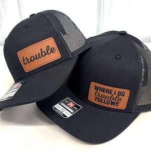 Può includere: Due cappellini da camionista neri con toppe in pelle marrone. Un cappellino recita "trouble" in corsivo. L'altro cappellino recita "WHERE I GO TROUBLE FOLLOWS" in grassetto. Entrambi hanno un retro in rete nera e una visiera curva.