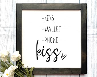 May include: A black framed sign with white background and black text that reads "Keys, Wallet, Phone, kiss" with a small heart symbol.