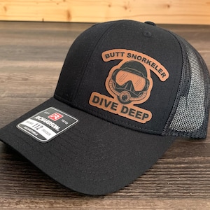 Puede incluir: Gorra de camionero negra con parte trasera de malla y parche de cuero. El parche presenta un gráfico de máscara de buceo con las palabras "BUTT SNORKELER" y "DIVE DEEP". La gorra tiene una correa ajustable y una etiqueta Richardson 112.
