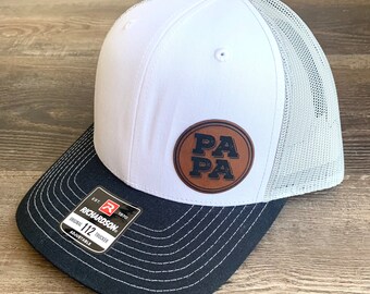 Könnte beinhalten: Weiße und schwarze Trucker-Kappe mit einem braunen Leder-Patch, auf dem in schwarzen Buchstaben "PA PA" steht.