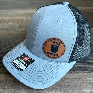 Könnte beinhalten: Eine graue und schwarze Trucker-Kappe mit einem braunen Leder-Patch, auf dem "FREE RIDES" mit einem Bart-Symbol steht. Die Kappe ist verstellbar.