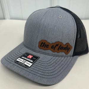 May include: A gray and black trucker hat with a brown leather patch that says "the ol' lady". The hat is made by Richardson 112 and is adjustable.