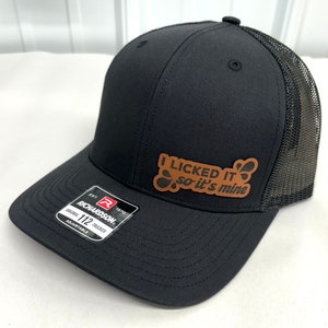 May include: Black and brown trucker hat with a brown leather patch that says "I licked it so it's mine".