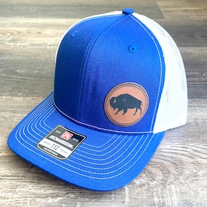 May include: A royal blue and white trucker hat with a brown leather patch featuring a silhouette of a buffalo. The hat is adjustable and has the text "Richardson 112 Trucker Adjustable" on the label.