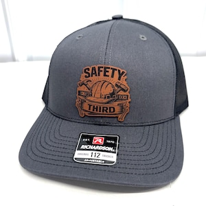 May include: A charcoal gray trucker hat with a black mesh back and a tan leather patch on the front. The patch features a graphic of a hard hat, hammer, and wrench with the text SAFETY THIRD. A black Richardson 112 sticker is on the brim.