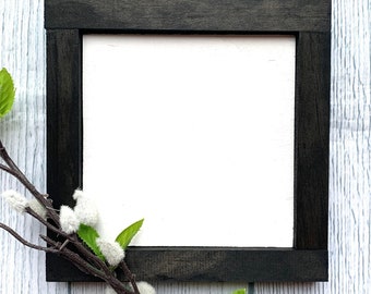 May include: A black wooden frame with a white background. The frame is on a white wood background with a sprig of green leaves and white flowers.