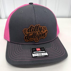 Puede incluir: Una gorra de camionero gris carbón con paneles traseros de malla rosa brillante y costuras rosas en la visera. Un parche de cuero marrón en la parte delantera presenta el texto negro 'dilly dallyin'. Una pegatina negra en la visera dice 'EST. 1970 RICHARDSON ORIGINAL 112 TRUCKER ADJUSTABLE'.