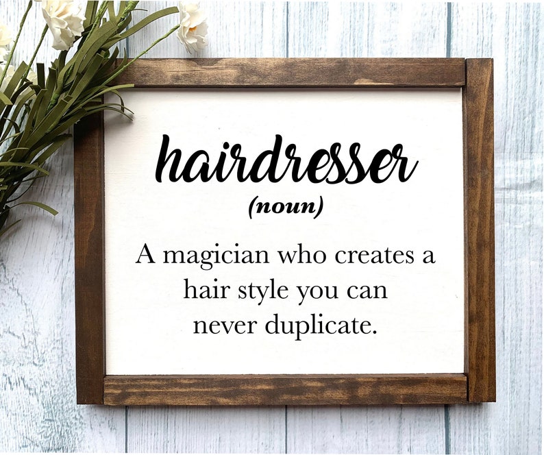 May include: A framed wooden sign with the word "hairdresser" in black script. Below, the definition reads: "A magician who creates a hair style you can never duplicate." The frame is dark brown, and the background is white.