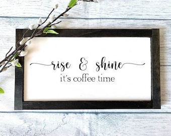 May include: A black framed sign with white background and the text "rise & shine it's coffee time" in black cursive font.
