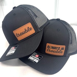 Puede incluir: Dos gorras de camionero negras con la parte trasera de malla. Una gorra tiene un parche de cuero marrón con la palabra "trouble". La otra tiene un parche de cuero marrón con las palabras "ALWAYS IN trouble".