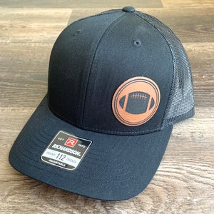 May include: Black trucker hat with a brown leather patch featuring a football design. The hat has a black mesh back and a black front with the text "EST. 1970 RICHARDSON. ORIGINAL 112 TRUCKER ADJUSTABLE" on a red tag.