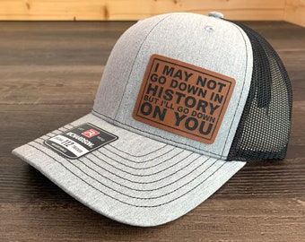 Könnte beinhalten: Eine graue und schwarze Trucker-Kappe mit einem braunen Leder-Patch, auf dem steht: "Ich werde vielleicht nicht in die Geschichte eingehen, aber ich werde dich runterziehen".
