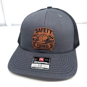 May include: A charcoal gray trucker hat with a black mesh back and a tan leather patch on the front. The patch features a graphic of a hard hat, hammer, and wrench with the text SAFETY THIRD. A black Richardson 112 sticker is on the brim.