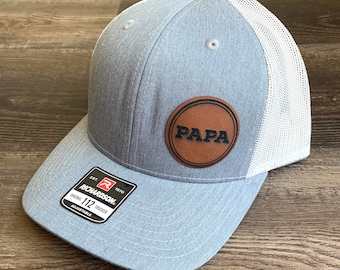 Könnte beinhalten: Eine graue und weiße Trucker-Kappe mit einem braunen Leder-Patch, auf dem in schwarzen Buchstaben "PAPA" steht. Die Kappe ist verstellbar und hat ein Richardson 112-Etikett.