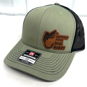 May include: A green and black trucker hat with a brown leather patch that says "Support Your Local Hookers" and a fish graphic. The hat is adjustable and has a Richardson logo.