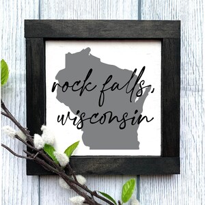 May include: A black framed sign with a white background and the text "rock falls, wisconsin" in black cursive font. The state of Wisconsin is outlined in gray.