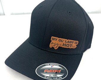 Peut inclure: Casquette de baseball noire avec un patch en cuir marron qui dit "MY OL' LADY IS PsycHOTIC". La casquette a un logo Flexfit et le texte "THE ORIGINAL FLEXFIT XL/XXL IT FITS YOUR LIFE".