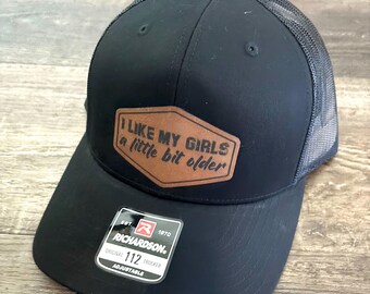 Peut inclure: Casquette de camionneur noire avec un patch en cuir marron qui dit "J'aime mes filles un peu plus âgées". La casquette est réglable et porte le logo Richardson.