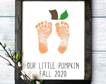 May include: A white wooden sign with a black frame, featuring a pair of orange baby footprints in the shape of a pumpkin with a green stem. The text "Our Little Pumpkin Fall 2020" is printed below the footprints.