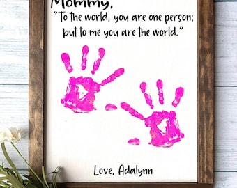 May include: A white wooden sign with a brown frame, featuring pink handprints and the text "Mommy, "To the world, you are one person; but to me you are the world." Love, Adalynn"