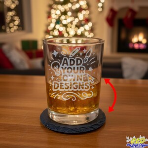May include: A clear glass tumbler with the text "ADD YOUR OWN DESIGNS" etched on the front, filled with a golden liquid. The glass sits on a round, black coaster. The background features a Christmas tree and fireplace.