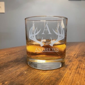 May include: A clear glass rocks glass with a frosted design of antlers and the letter A. The glass is filled with amber liquid and ice cubes. The name "ANDREWS" is frosted on the bottom of the glass.