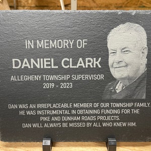 K&ouml;nnte beinhalten: Eine schwarze Schiefertafel mit einer wei&szlig;en, gravierten Inschrift zum Gedenken an Daniel Clark, Supervisor der Gemeinde Allegheny, 2019-2023. Die Inschrift enth&auml;lt ein Foto von Daniel Clark und eine Nachricht &uuml;ber seine Beitr&auml;ge zur Gemeinde.