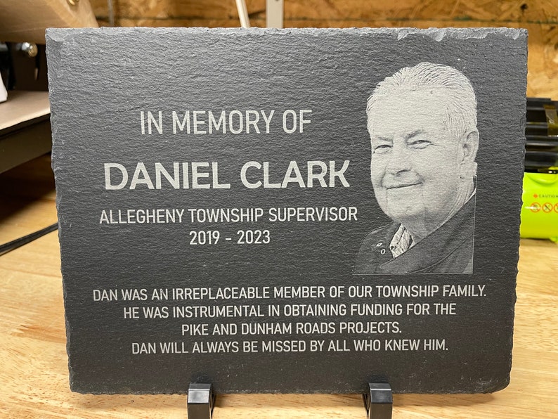 K&ouml;nnte beinhalten: Eine schwarze Schiefertafel mit einer wei&szlig;en, gravierten Inschrift zum Gedenken an Daniel Clark, Supervisor der Gemeinde Allegheny, 2019-2023. Die Inschrift enth&auml;lt ein Foto von Daniel Clark und eine Nachricht &uuml;ber seine Beitr&auml;ge zur Gemeinde.